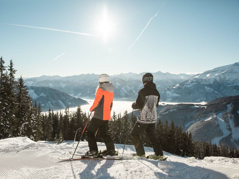 Zwei Skifahrer stehen auf einem Berg und blicken auf die schneebedeckte Landschaft. | © Schmittenhöhebahn AG/Punkt und Komma