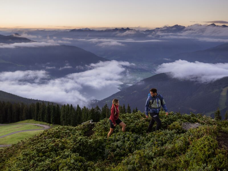 Zwei Wanderer erkunden die grüne Berglandschaft mit Wolken und Bergen im Hintergrund. | © Schmittenhöhebahn AG/mr-offenblende.at