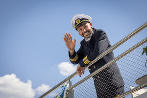 Ein Kapitän in Uniform winkt von einem Geländer auf einem Schiff. | © Nikolaus Faistauer Photography