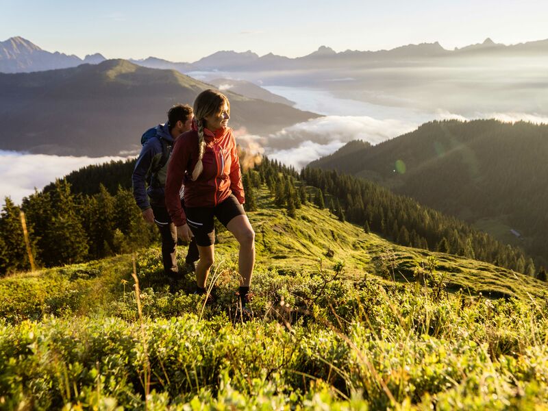 Zwei Personen wandern auf einem Hügel in den Bergen, umgeben von Nebel und einem atemberaubenden Ausblick. | © Schmittenhöhebahn AG/mr-offenblende.at