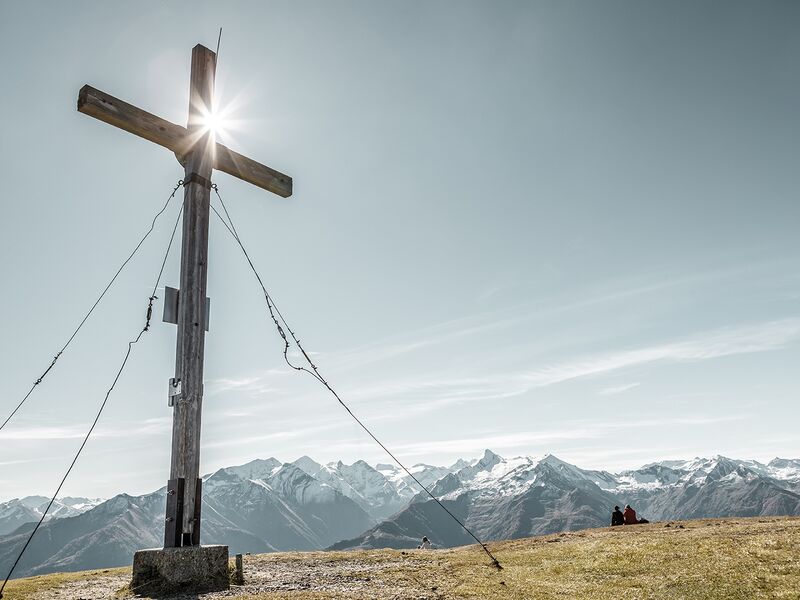 Holzkreuz mit Sonnenstrahlen und Blick auf die Berge. | © Schmittenhöhebahn AG/Christian Woeckinger