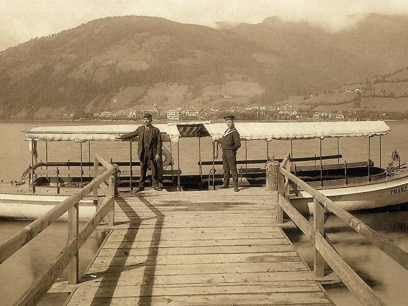 Zwei Männer stehen neben einem nostalgischen Boot am Zeller See, Berge im Hintergrund. | © Bezirksarchiv Pinzgau