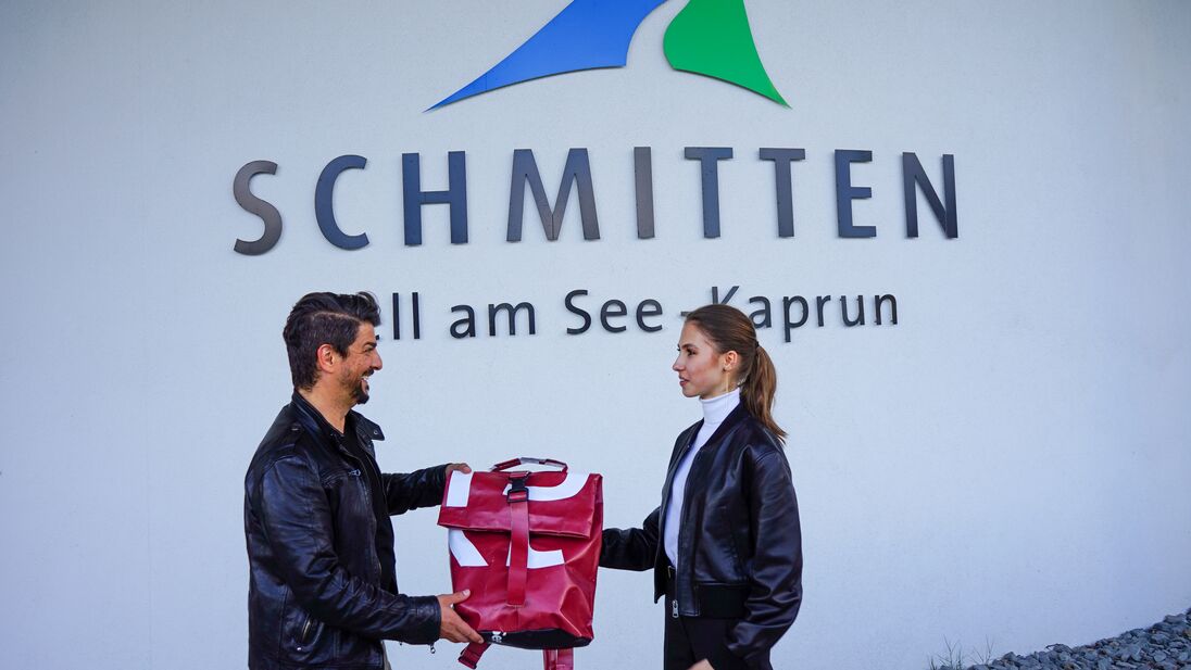 Zwei Personen bei einer Geschenkübergabe vor dem Schmitten-Logo in Zell am See. | © Schmittenhöhebahn AG