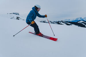 Person mit blauer Jacke und roten Ski auf der Piste vor einer schneebedeckten Berglandschaft.