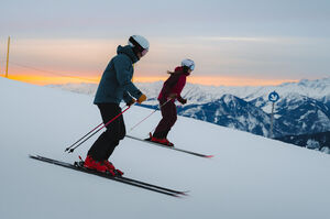 Zwei Skifahrer bei Sonnenaufgang auf einer Piste mit Bergen im Hintergrund.