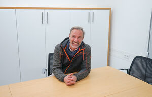 Ein Mann sitzt an einem Tisch und lächelt in die Kamera, vor einem weißen Schrank. | © Schmittenhöhebahn AG
