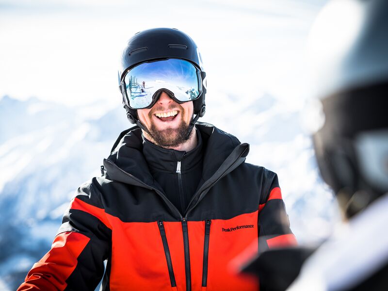 Lachender Mann im Skianzug mit Helm und Skibrille, schneebedeckte Berge im Hintergrund. | © Bründl Sports