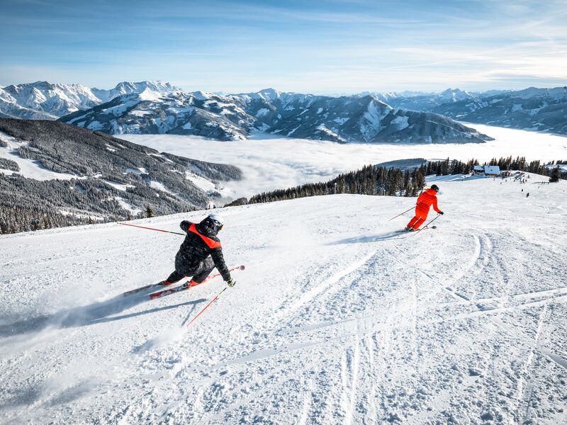 Zwei Skifahrer bei herrlichem Wetter auf einer verschneiten Piste mit Alpenpanorama. | © Bründl Sports