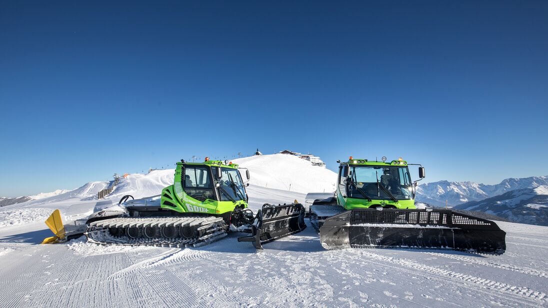 Zwei grüne Pistenraupen präparieren eine Skipiste auf einem Berg bei klarem Himmel. | © Nikolaus Faistauer Photography