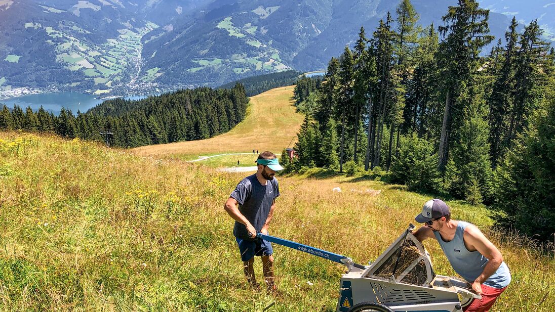 Zwei Männer mit einer Maschine auf einer Almwiese vor Bergkulisse. | © Schmittenhöhebahn AG