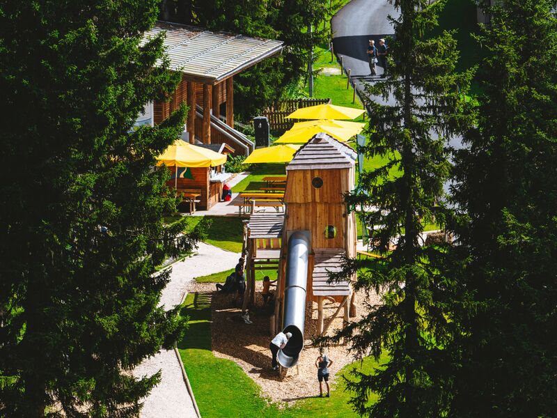 Ein Waldspielplatz mit Rutsche, Bäumen und gelben Sonnenschirmen. | © Alpine Vision
